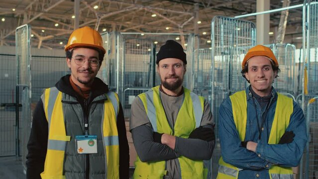 Medium portrait of three diverse male warehouse workers in safety uniform and hardhats posing in storage room and looking at camera