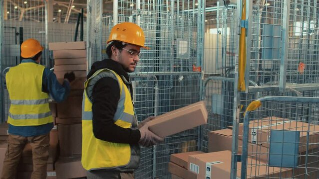 Medium side shot of Biracial warehouse worker in glasses and hardhat segregating boxes with stock goods on cart indoors