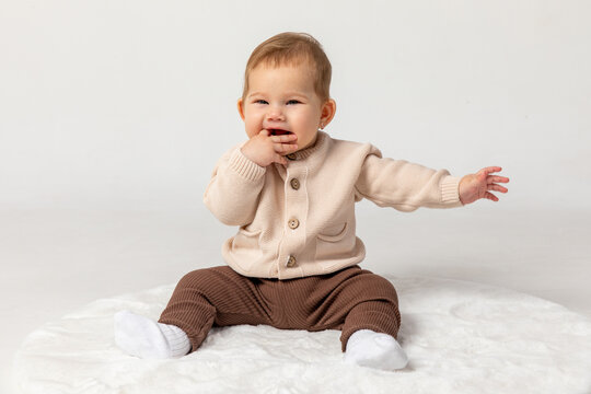 Happy smiling adorable newborn baby girl in beige and brown suit sitting on light white home carpet on white background. 6 to 7 months old development. advertising of toys and goods for children