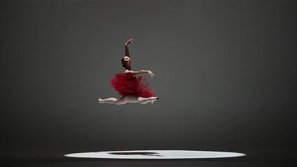 A ballerina in a red tutu and pointe shoes during a twine jump. A young woman dancing elements of classical ballet in a dark studio with overhead light. © kinomaster