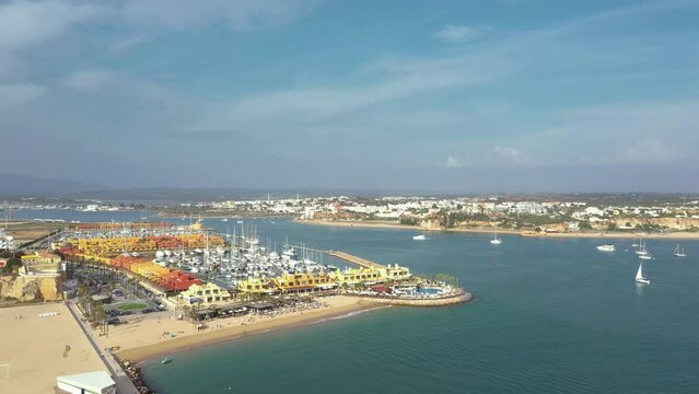 Cinematic aerial perspective of Portimao Marina. Luxury yacht docked in the port. Arade river in the middle. Ferragudo city in background. Drone backwards ascending. Famous travel destination