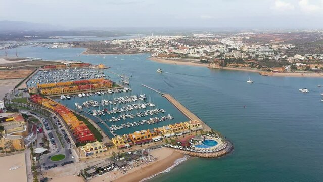 Cinematic aerial perspective of Portimao Marina. Luxury yacht docked in the port. Arade river in the middle. Ferragudo city in background. Drone descending. Famous travel destination