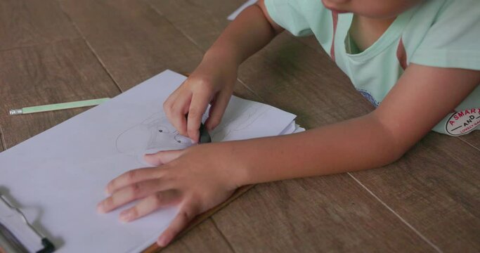 Asian child with black hair with a smiling face lying on the floor of his house drawing picture of a girl with braids. using his imagination creating art. Concept of children, learning, creativity. 4k