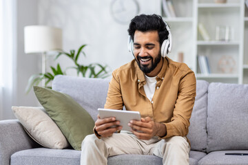 Indian young man sitting on the couch at home wearing headphones and using a tablet with a smile.
