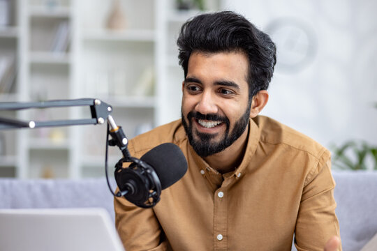 Close-up photo of a young Indian man smiling sitting on the couch at home in front of a microphone stand and talking.