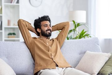 Happy Indian young man sitting relaxed on sofa at home with hands behind head and resting, smiling contentedly.