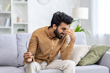 Indian young man sitting on sofa at home, holding glasses and laughing heartily with hand covering...