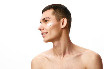 Handsome young man with short hair applying face moisturizing cream against white studio...