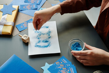 Hand of young unrecognizable woman sprinkling blue powder paint on paper sheet with outlines of...