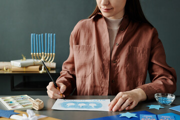 Cropped shot of young woman in brown shirt writing down Hanukkah wish on handmade greeting card...