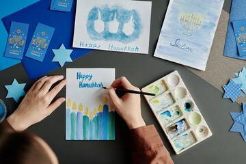 Hands of young creative woman with paintbrush and blue watercolor writing down Happy Hanukkah on...