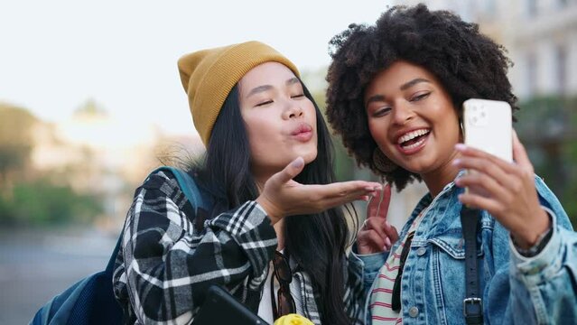 Cheerful multicultural female students with backpacks take selfies with cell phone and show different gestures on the street