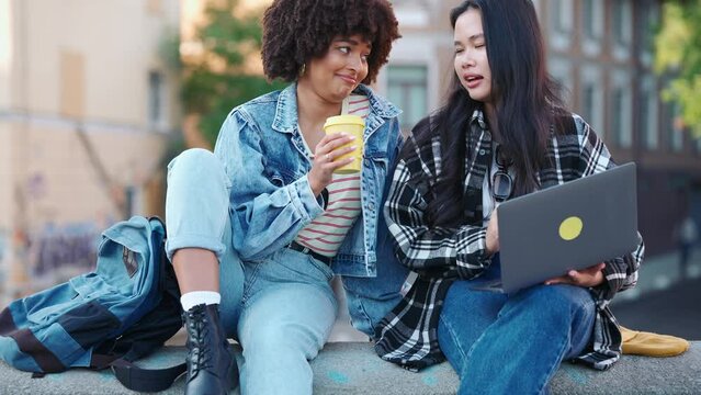 Cheerful multicultural female friends talking and looking at laptop on street in fall