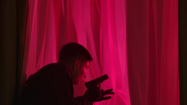 A male criminal with a gun sneaks up to a window in a dark room and looks out at the flashing police lights, close up. Male criminal walks away, hoping to evade police.