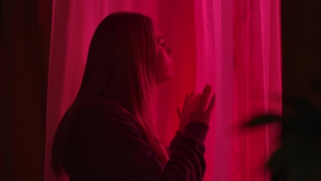 A concerned woman looks out the window at the flashing red and blue lights of the emergency services, police. The woman folds her hands in a prayer gesture and says a prayer, close up.