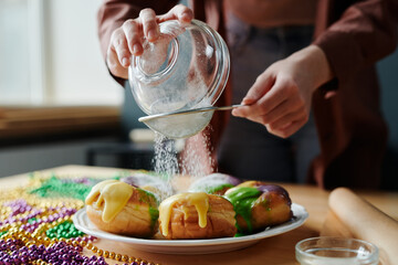 Hands of young unrecognizable woman sieving sugar powder on top of appetizing homemade donuts on plate while preparing for Mardi Gras - obrazy, fototapety, plakaty