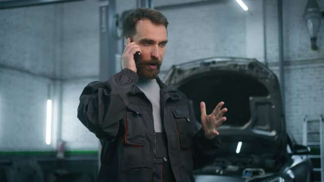 Annoyed serious Caucasian adult 30s man in uniform auto mechanic engineer talking mobile phone in repair car service male worker of technical industry communicate speak smartphone at vehicle workshop