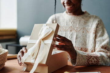 Hands of happy young African American woman in sweater opening giftbox with tied knot of white...