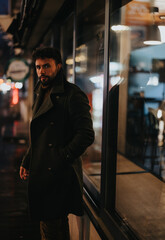 A stylish, good looking businessman standing on a sidewalk in a busy city at night. He is smoking a cigarette while waiting for a taxi to take him home after work on a rainy day.