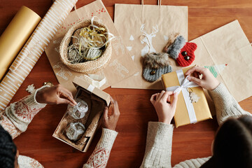 Above view of two young intercultural women sitting by table while one of them opening giftbox with two silver decorations for xmas tree