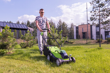 A young woman with a green lawn mower mows the lawn - obrazy, fototapety, plakaty