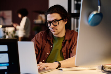 Young serious designer in eyeglasses looking at laptop screen and typing on keypad while working...
