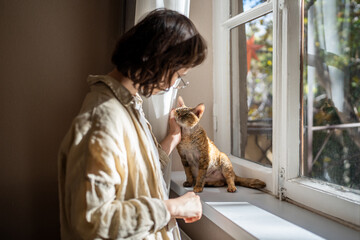 Tender loving teenager caressing, stroking adorable breed cat Devon Rex sitting on windowsill,...
