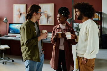 Group of happy intercultural designers having coffee and discussing working moments at break while standing in coworking space