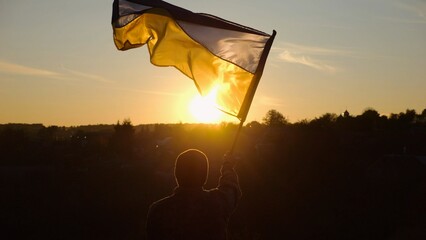 Young man in military uniform waving flag of Ukraine against beautiful sunset at background. Male...
