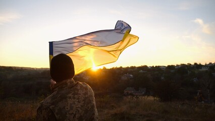 Young man in military uniform waving flag of Ukraine against beautiful sunset at background. Male...