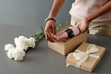 Young unrecognizable woman putting bottle of champagne in giftbox while packing present for mother...