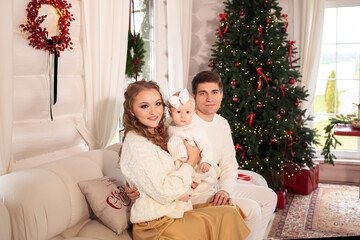 young parents with a little daughter in their arms in a beautiful room decorated for Christmas are sitting on a white sofa. father and mother playing with daughter on the sofa