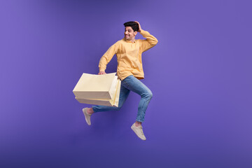 Full length photo of handsome young guy jumping run hold shopping bags dressed stylish yellow garment isolated on purple color background - obrazy, fototapety, plakaty