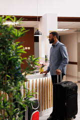 Young African American man with suitcase standing at reception area, talking pleasantly with front...