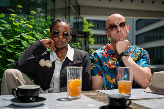 Fashionable mid-age man carrying a colorful hawaiian shirt and actractive young woman of color dressed in black sit at a bar table with drinks posing with serious expression