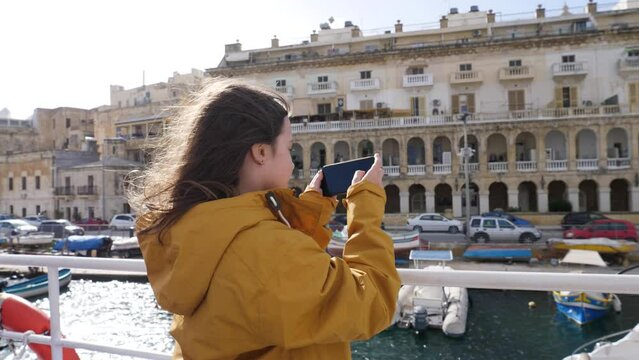 Kid girl tourist with smartphone on ferry boat takes picture of beautiful Malta architecture along a bay waterline