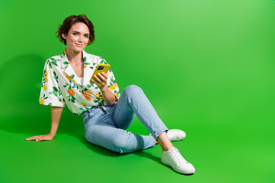 Full length photo of glad young girl sitting floor wear denim jeans trendy shirt surfing internet phone isolated on green color background