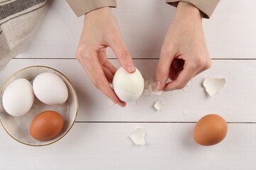 Woman peeling boiled egg at white wooden table, top view
