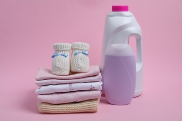 Laundry detergents, stack of baby clothes and small booties on pink background