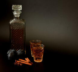 Plum liqueur in a crystal decanter and two glasses on a black background, next to ripe fruits and...