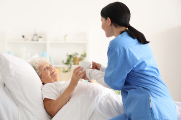 Senior woman taking cup of tea from nurse in bedroom