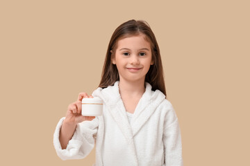 Cute little happy girl in bathrobe with jar of cream on beige background