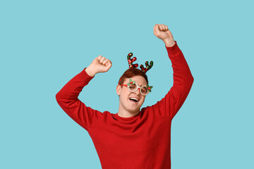 Cheerful young man in warm sweater, reindeer headband and novelty glasses on blue background