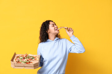 Beautiful young happy African-American woman eating delicious pizza on yellow background