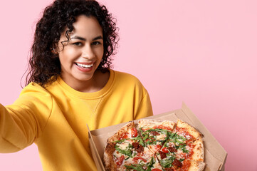 Beautiful young happy African-American woman with delicious pizza taking selfie on pink background - obrazy, fototapety, plakaty