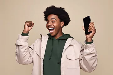 Tuinposter happy african american teenager ethnic cool guy student winner holding cell phone winning money online cellphone using mobile app celebrating gre result yes isolated beige background © sandra