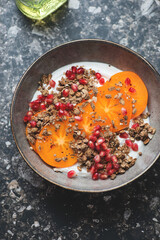 Plate of greek yogurt topped with granola, sliced persimmon and pomegranate seeds, vertical shot on a dark-brown granite background, elevated view