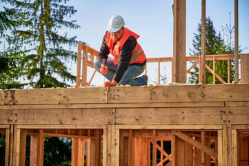 Carpenter constructing two-story wooden frame house near forest. Bearded man hammering nails into the structure, wearing protective helmet, construction vest. Concept of modern ecological construction