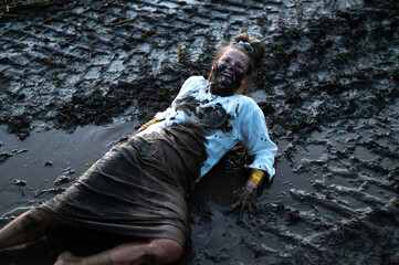 Woman with a dirty black face and fancy copper bracelets is stuck in a mud puddle on a field