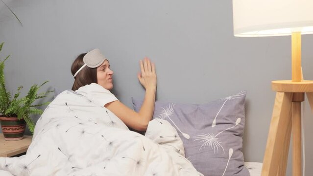 Unhappy young woman in sleeping mask on her head suffering and disturbed by noisy neighbors calling on smartphone knocking to wall trying to falling sleep in bed at home at night.